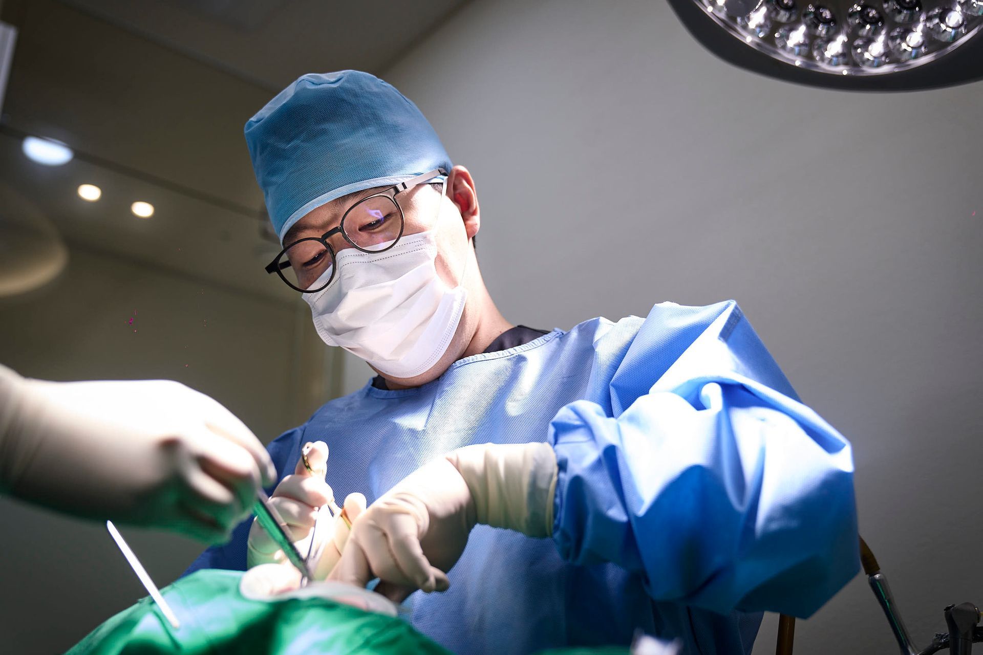Surgeon in operating room, performing surgery on a patient, wearing blue scrubs, mask, and gloves.