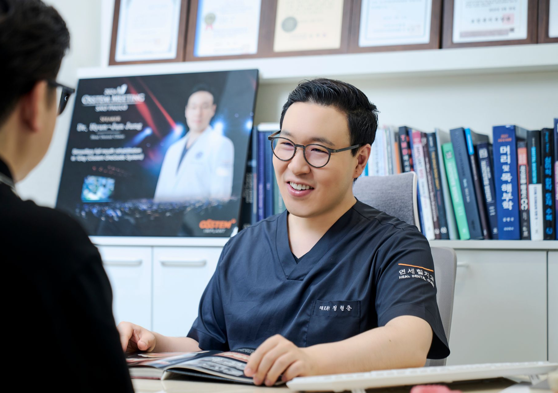Man in scrubs consults with a patient, smiling. Office setting, books and diplomas visible.