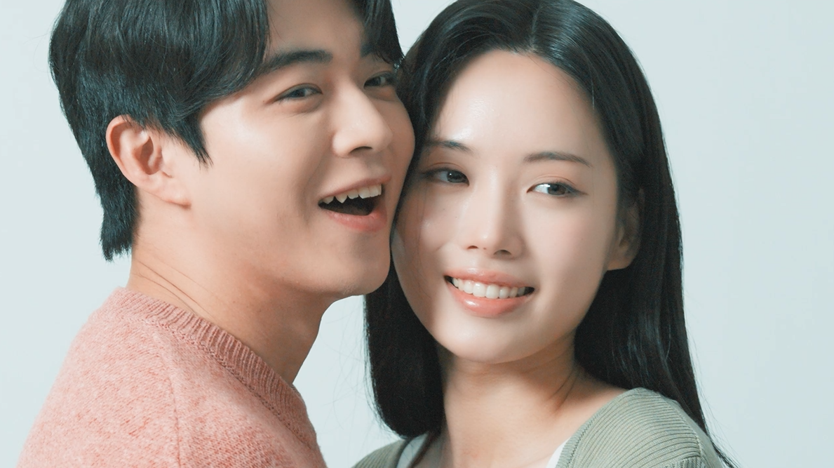 Man and woman smiling, cheek-to-cheek. Man wears pink, woman wears green, both have dark hair. White background.