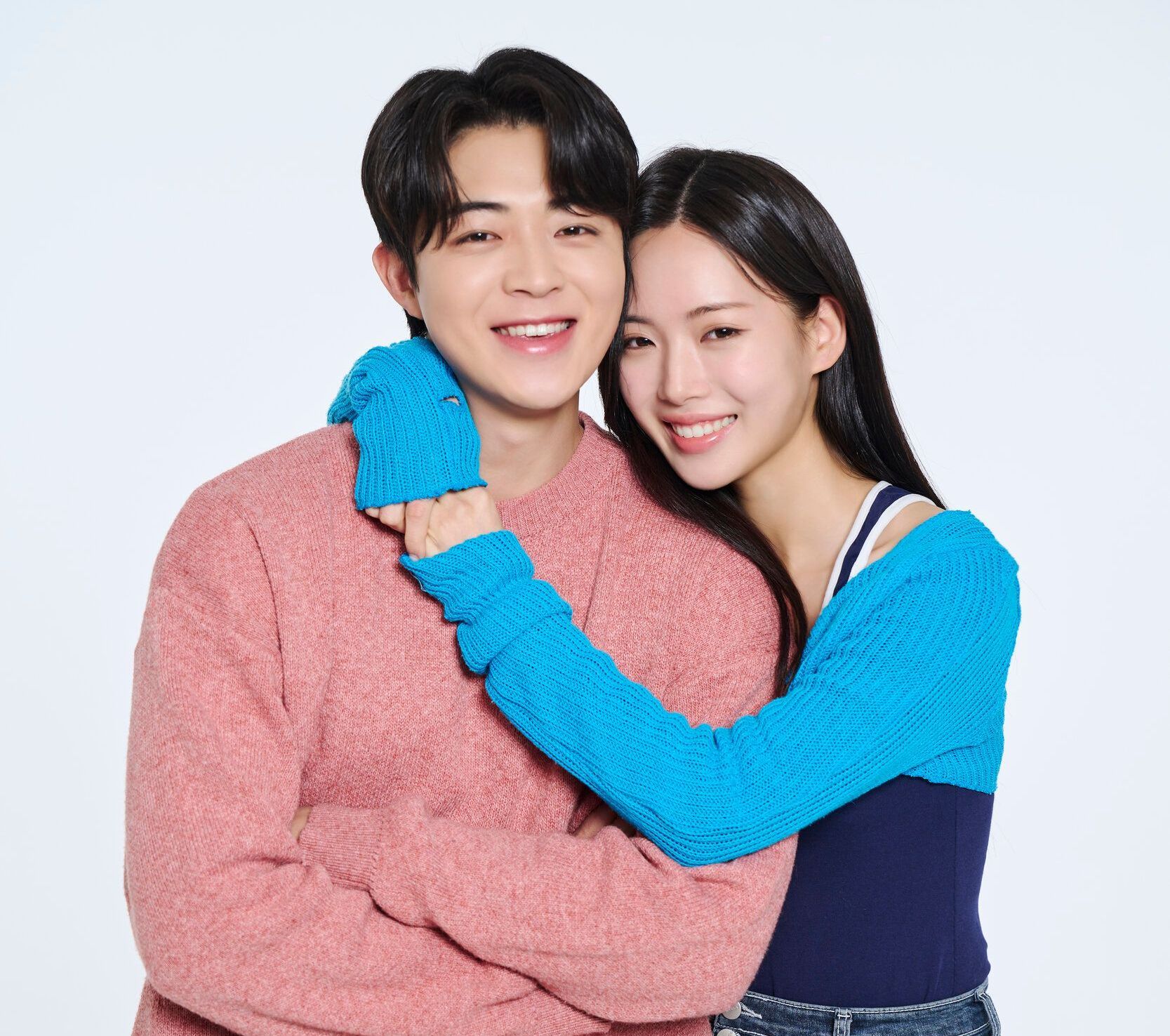 Smiling couple hugging against a white background; man in pink sweater, woman in blue sweater.