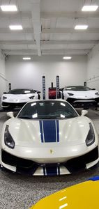 A white sports car with blue racing stripes parked in a bright garage with two other luxury cars in the background.