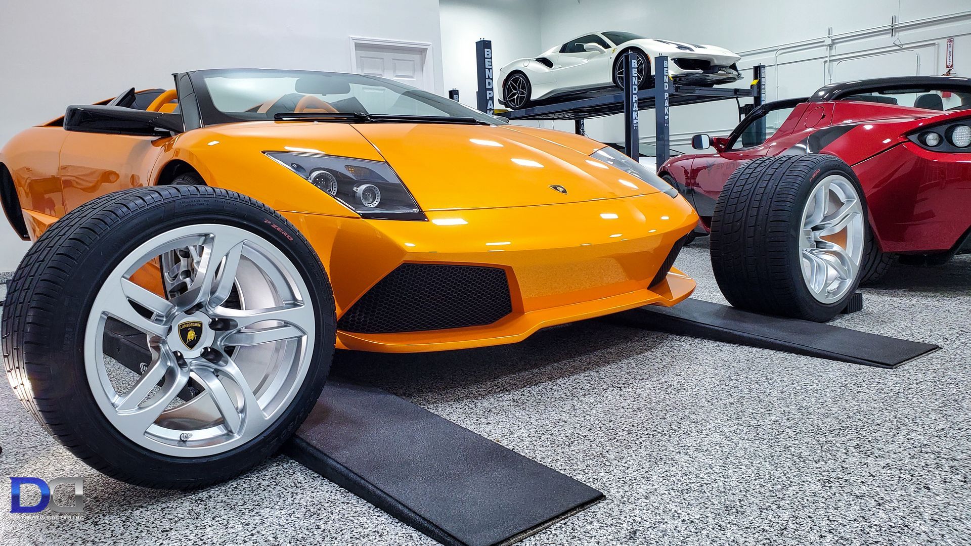 An orange Lamborghini convertible parked on shop ramps in a garage with other luxury cars nearby.