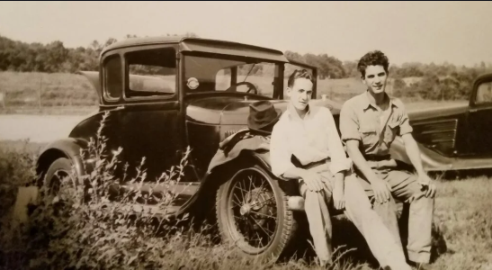 Two men are sitting next to an old car in a field.