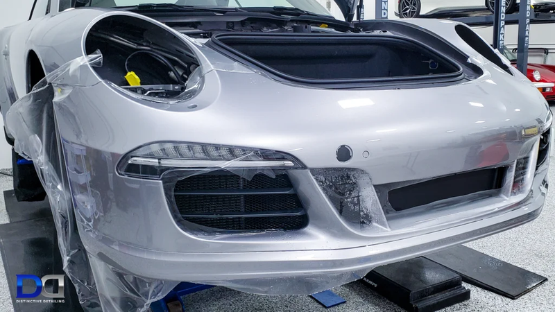 Silver Porsche's front end, with hood open, undergoing detailing. Plastic wrap covers bumper.