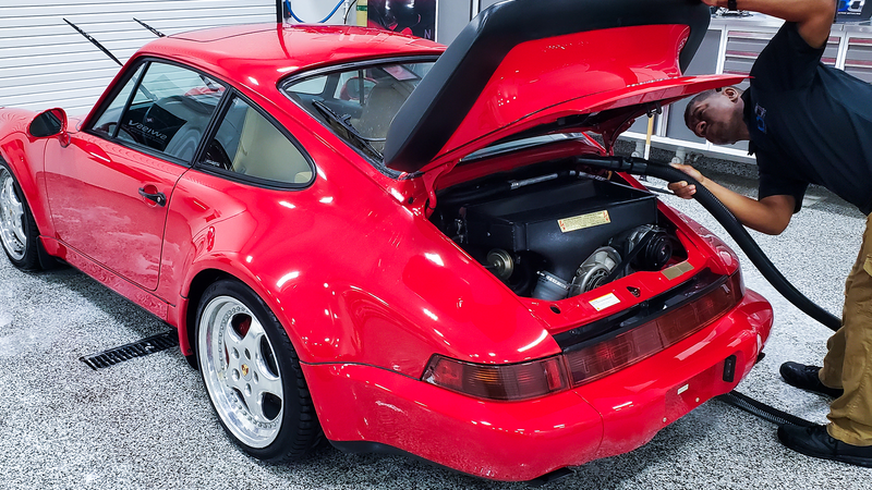 A person vacuums the engine bay of a bright red Porsche 911 sports car.