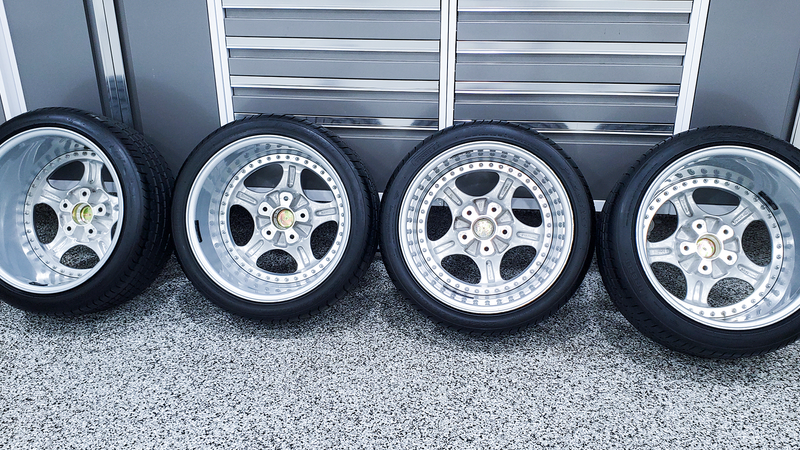 Four silver car wheels with black tires on a gray speckled surface, near a gray wall.