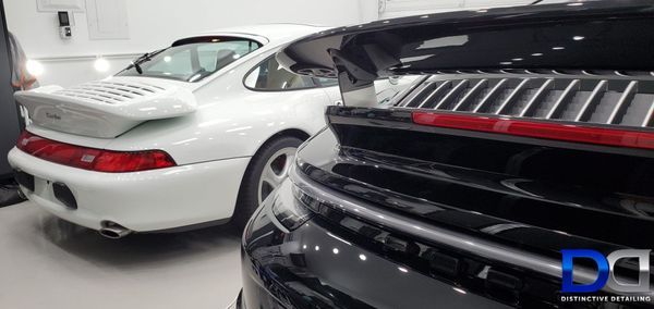 Two Porsche 911s, white and black, with prominent rear spoilers, in a bright interior space.