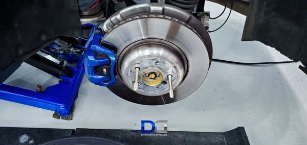 A car's rear brake assembly. The blue brake caliper surrounds the rotor, with a blue jack stand on the left.