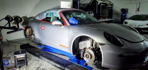 A silver Porsche on a lift in a garage, with one wheel removed for repair or maintenance.