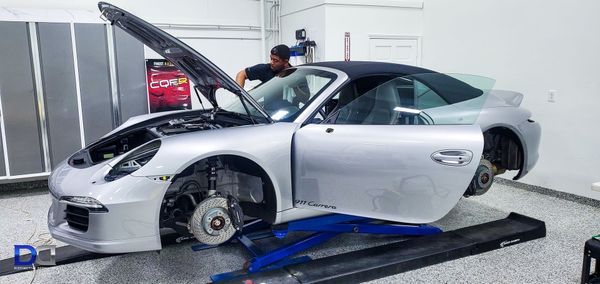 Mechanic working on a silver Porsche in a garage with the hood and door open.