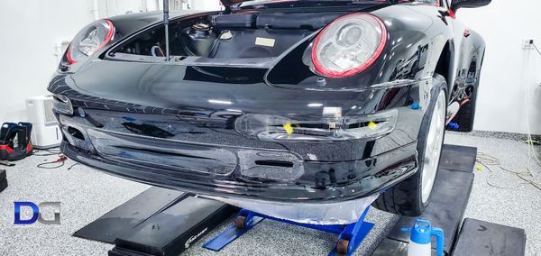 Black Porsche on a lift in a garage with its hood open. Front bumper is detached.