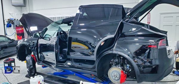 Black Tesla car being repaired in a workshop with open doors, the hood, and a mechanic working on it.