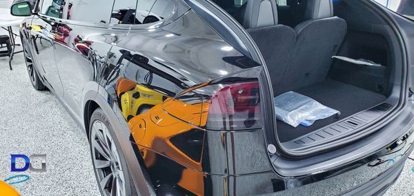 Black Tesla SUV with open trunk; a yellow and orange car are reflected on the body. Inside a bag.