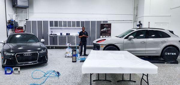 A man stands in a garage with two cars: a black Audi and a gray Porsche. There's a white table in the foreground.