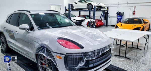 White SUV in a garage with other cars on lifts and on the ground; clear film covering parts of the vehicle.
