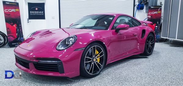 Pink Porsche 911 Turbo S with yellow calipers parked in a garage, with banners and equipment in the background.