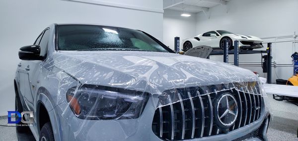 A light gray Mercedes SUV covered in protective film in a garage with a white sports car on a lift.