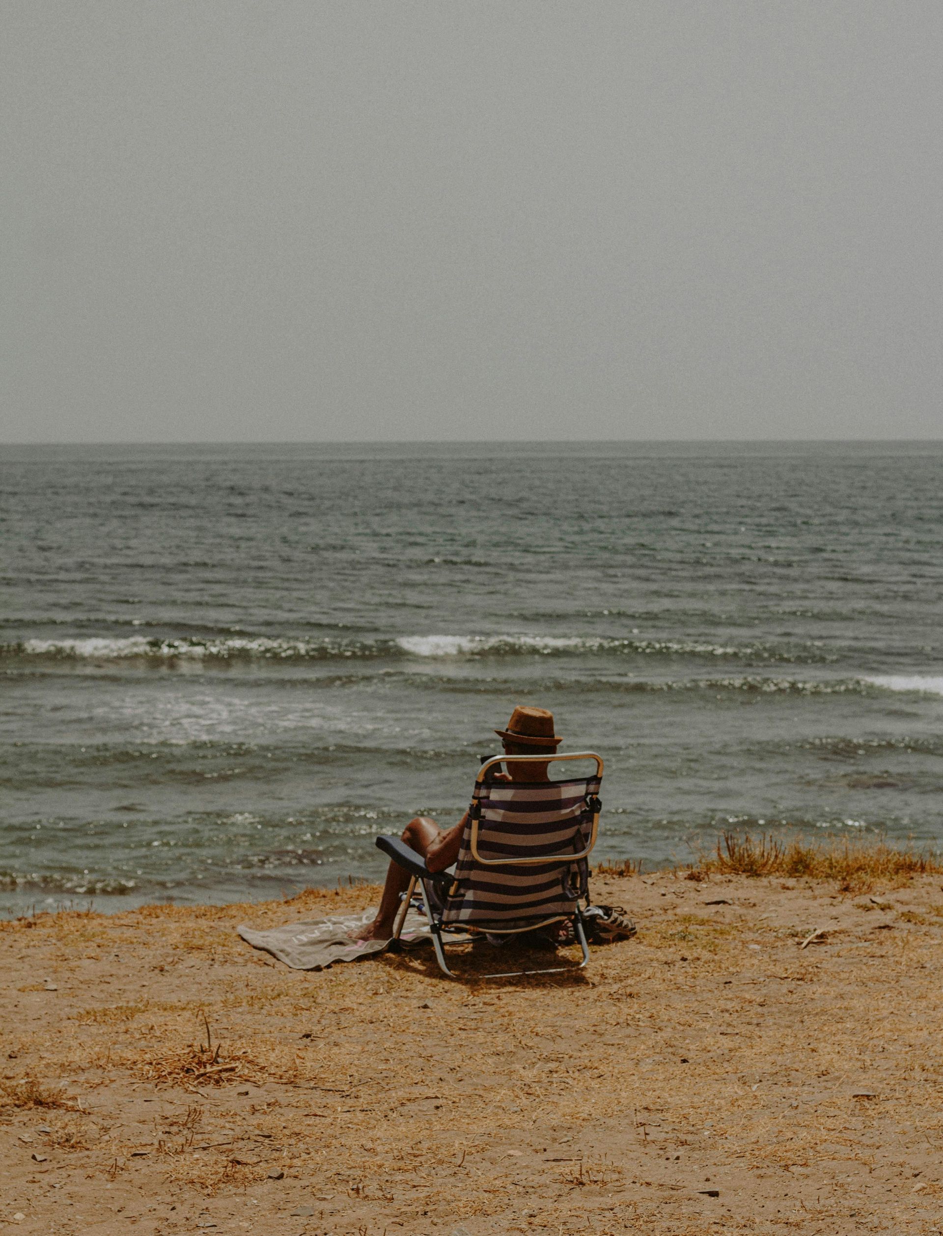 A man is sitting in a chair on the beach looking at the ocean.