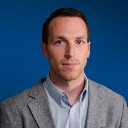 A man in a suit and blue shirt is standing in front of a blue background.