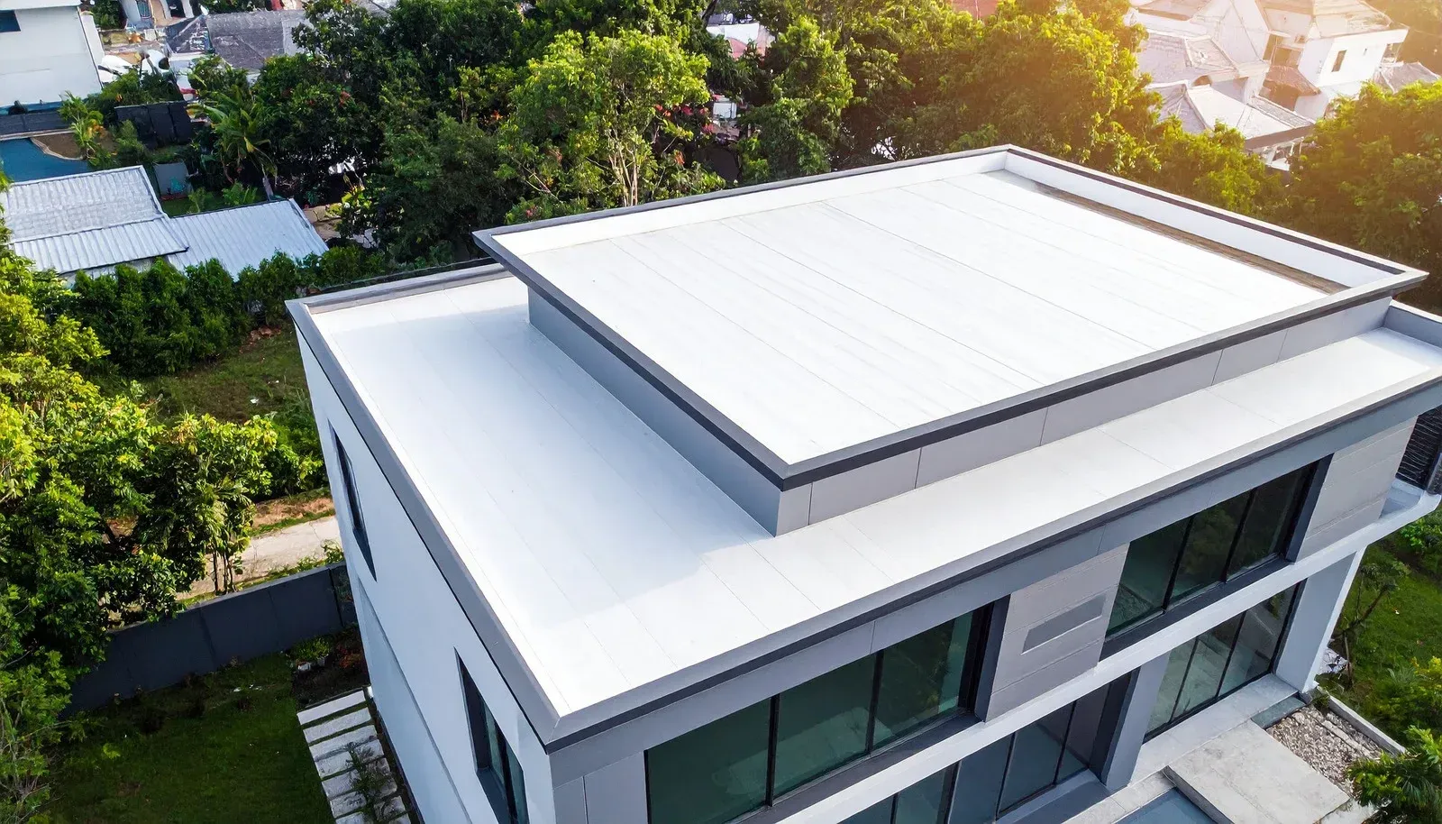 Modern two-story house with a flat white roof surrounded by lush green trees and other buildings.