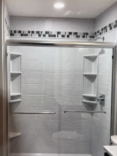 White and gray tiled shower with sliding glass doors and built-in shelves, accented with black mosaic tile.