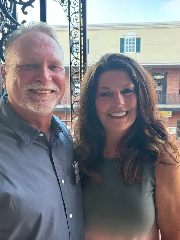 Man and woman smiling together outdoors. The man wears a gray shirt. The woman wears a green top.