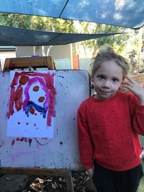 A little girl is standing in front of an easel with a painting on it.