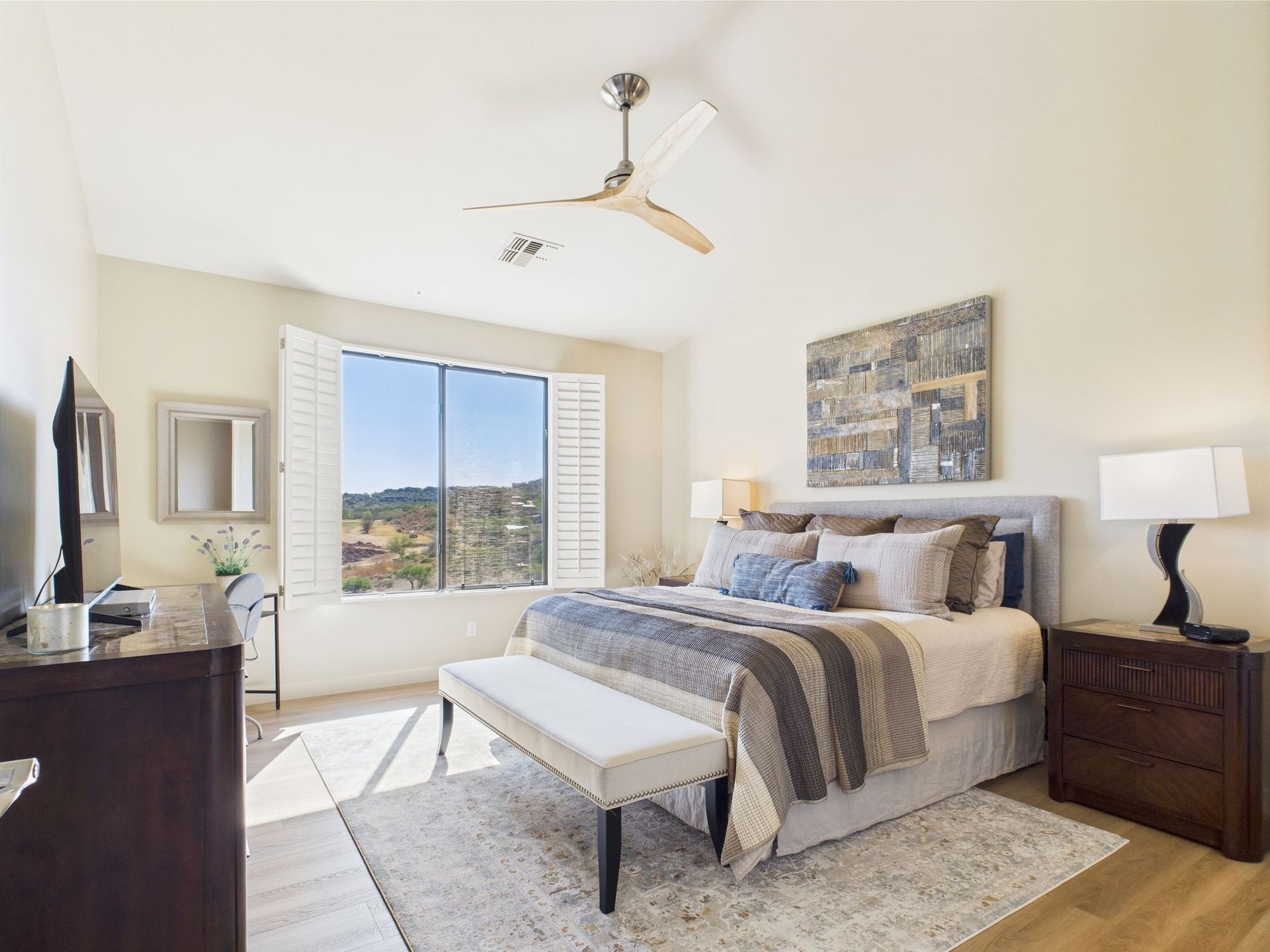 Bedroom with king bed, shutters, and bench. Light colors, sunny.