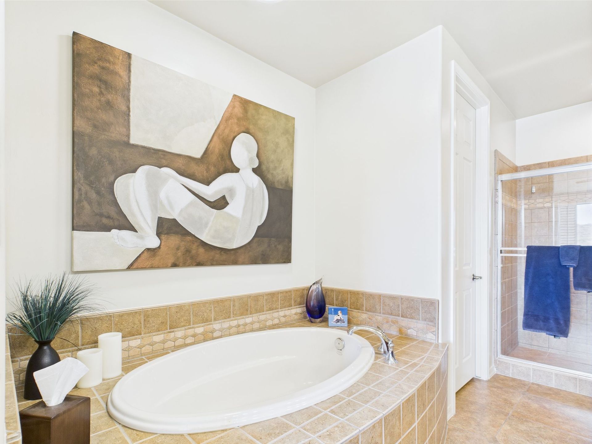 Bathroom with oval tub, beige tile, abstract art, white walls, glass shower, and blue towels.