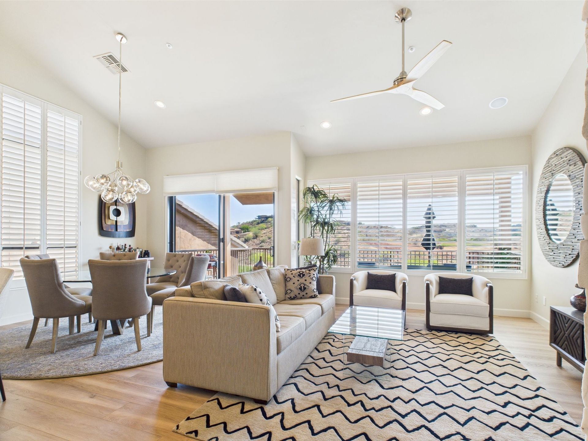 Bright, airy living room with beige sofa, white chairs, and patterned rug, near a dining area with a view.