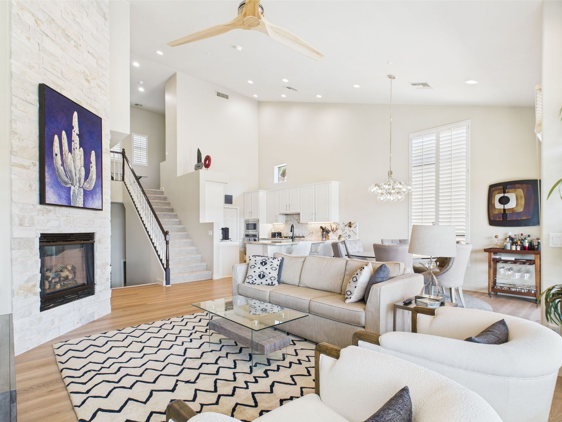 Living room with high ceilings, fireplace, stairs, and open kitchen. Beige, white, and black decor.