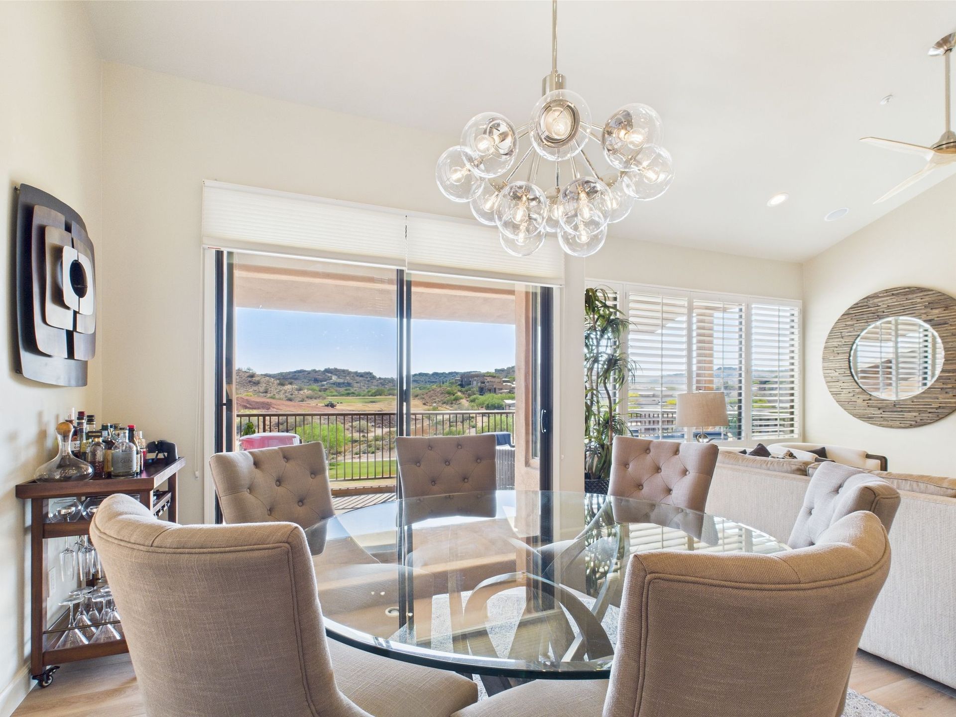 Elegant dining room with glass table, tufted chairs, and a chandelier, overlooking a scenic landscape.