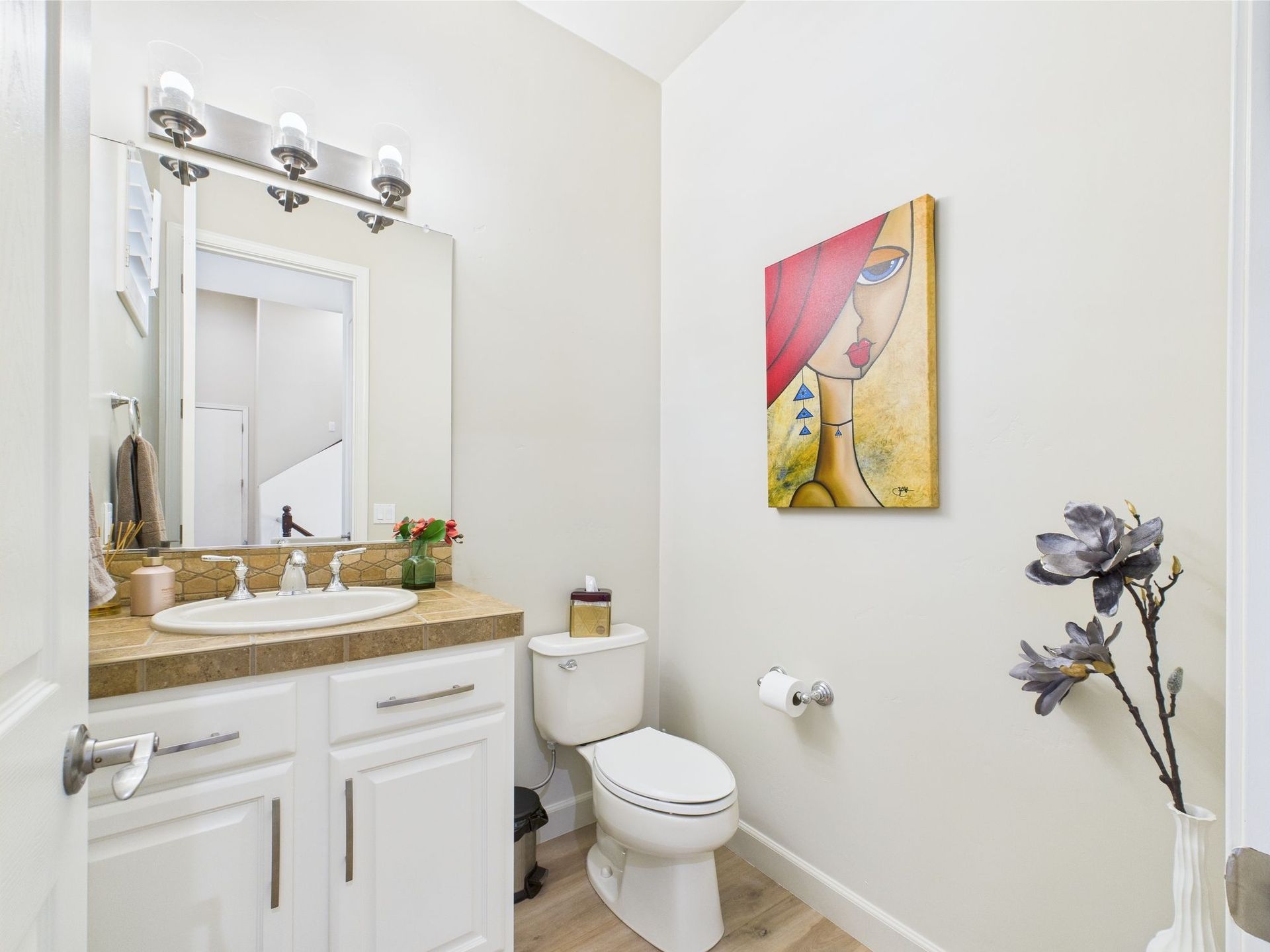 Small bathroom with a white toilet, vanity, and artwork; neutral walls, wood-look flooring.