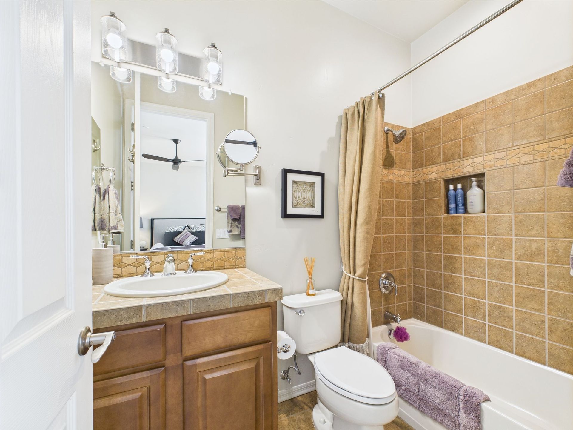 Bathroom with brown tile, beige shower curtain, vanity, toilet, and a glimpse into a bedroom.