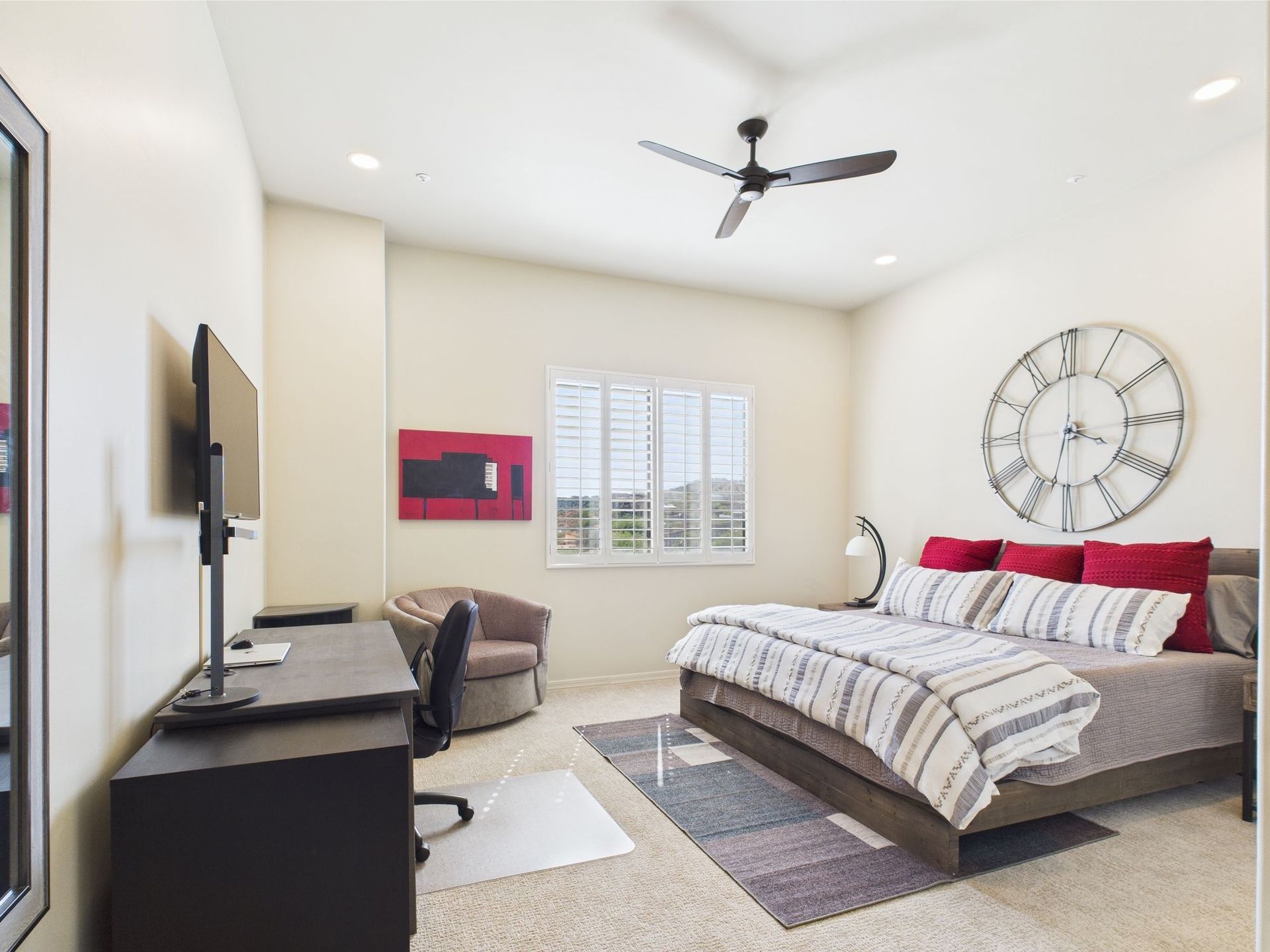 Bedroom with bed, desk, armchair, large clock, and window with blinds. Neutral color scheme.