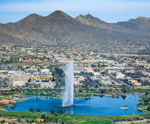 Cityscape with lake, fountain, buildings, and mountains in the background under a blue sky.