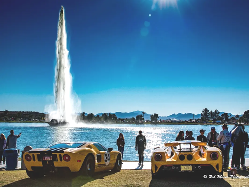 Two yellow Ford GTs parked by a lake with a tall fountain under a blue sky, people watching.
