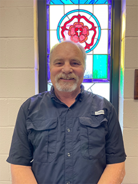 A man in a blue shirt is standing in front of a stained glass window.