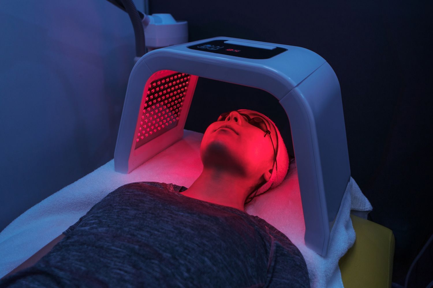 A woman is laying on a bed getting a red light treatment on her face.