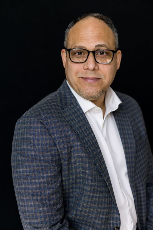 A man wearing glasses and a suit jacket is standing in front of a black background.