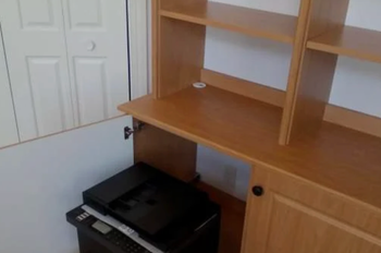 A printer is sitting under a wooden desk in a room.
