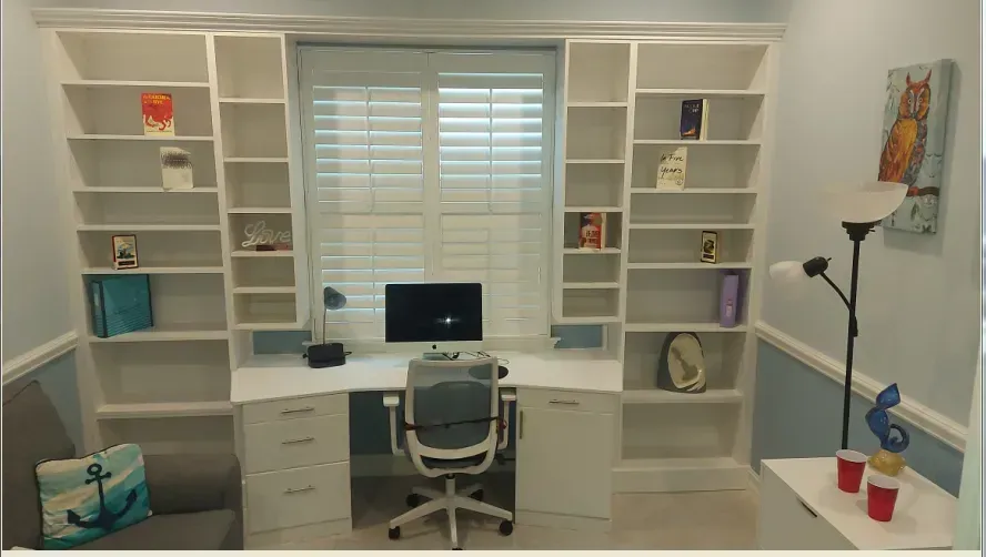 A room with a desk , chair , shelves and a window.