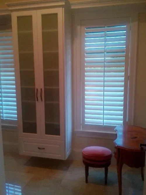A room with a desk , chair , ottoman , and shutters on the windows.