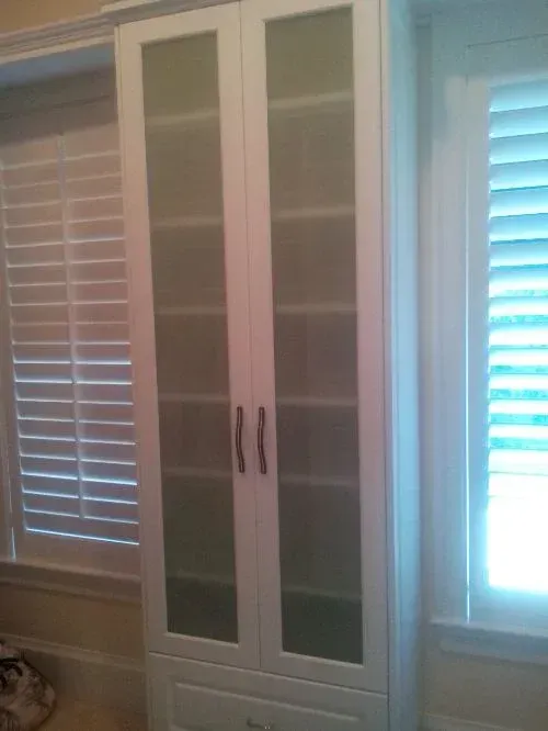 A white cabinet with glass doors is next to a window with blinds.