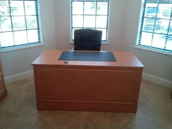 A wooden desk with a black chair in front of two windows
