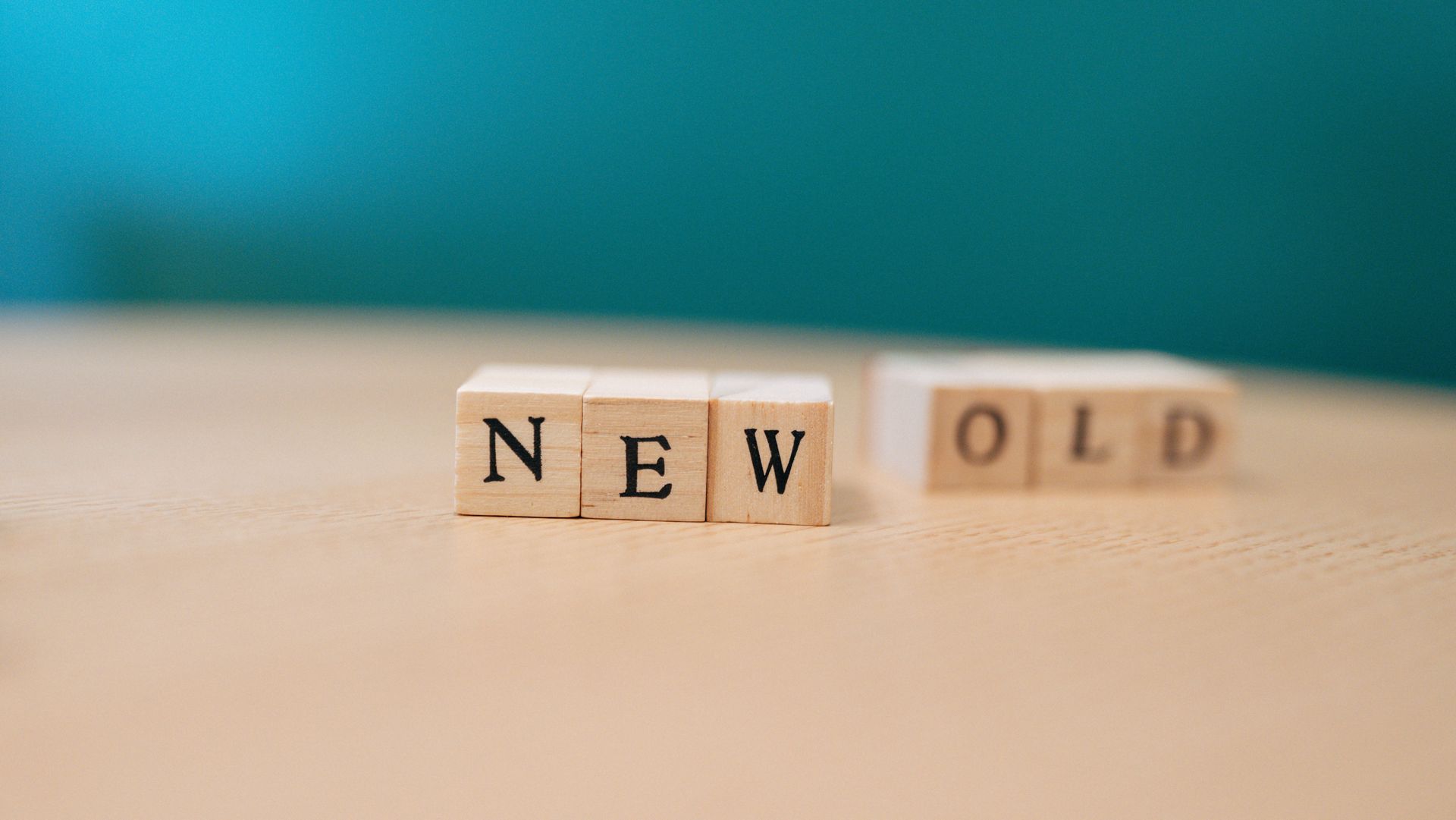 Wooden blocks spelling “NEW” beside “OLD” on a beige surface with teal background.