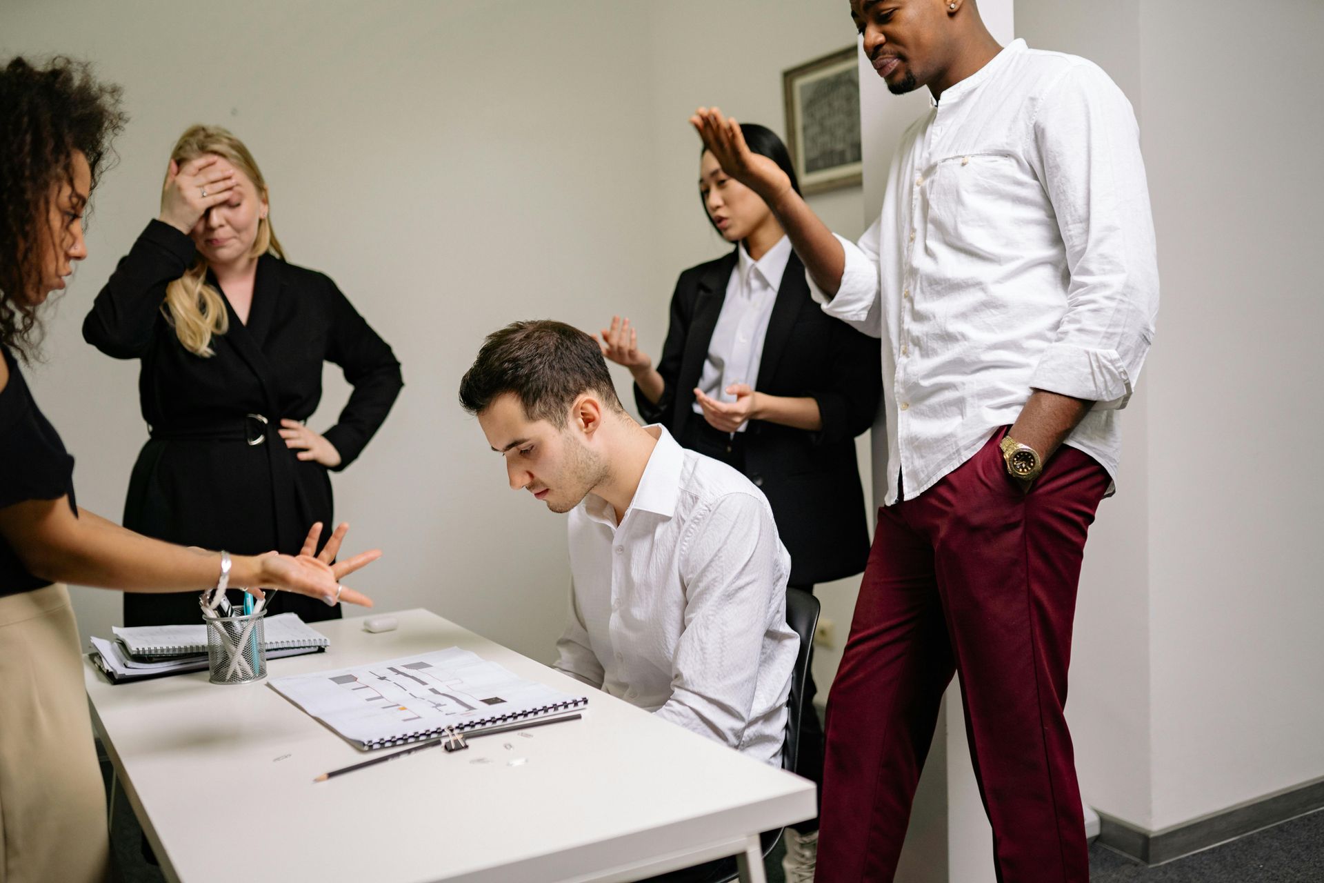 People in an office meeting, one man leaning over a table while others stand nearby with concerned expressions