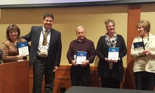 Five people holding certificates onstage, smiling. Indoors, neutral background.