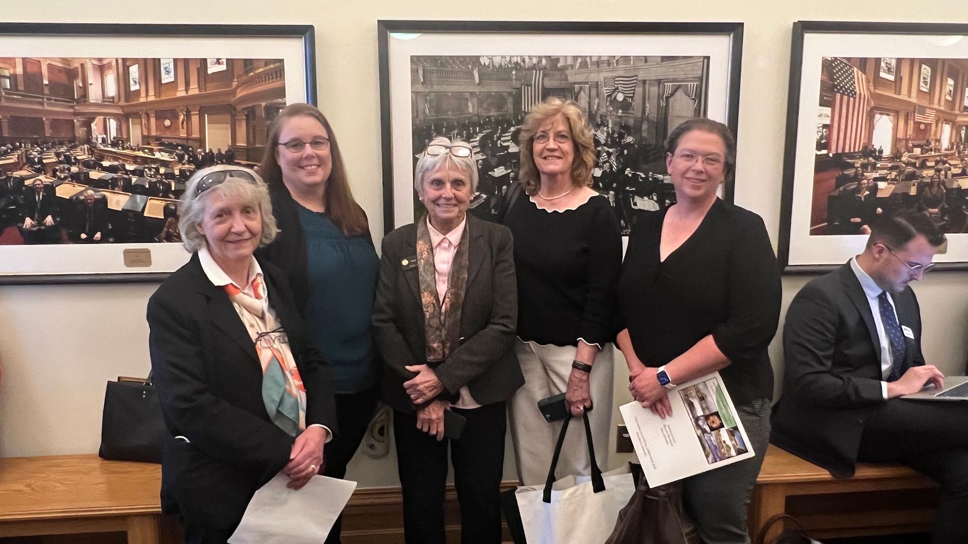 Five women posing in front of framed historical images in a building. One person is sitting on a bench to the right.