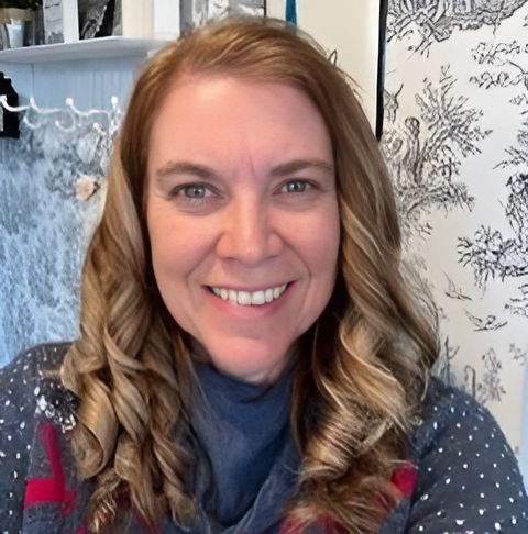 Woman with wavy blonde hair smiling at the camera in front of a patterned backdrop.